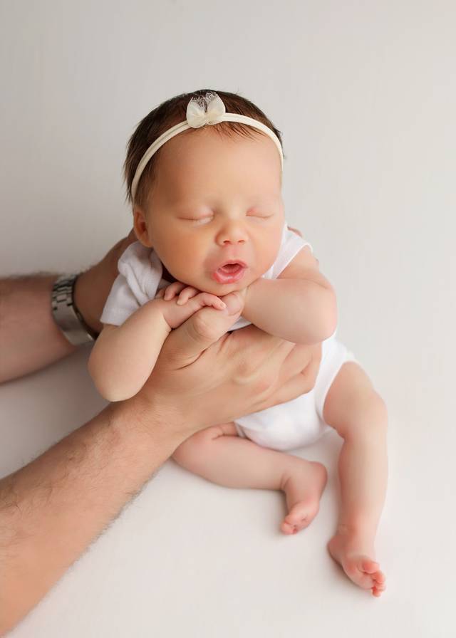 A newborn in a white onesie held in her father's hands.
