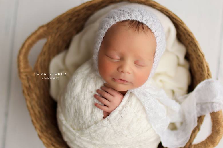 Newborn in a white knit bonnet, wrapped and asleep in a woven basket.