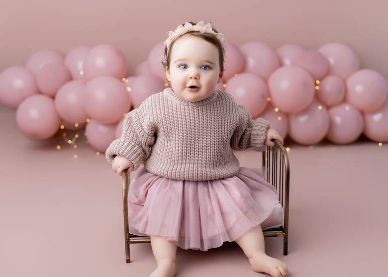 One year old in a tulle skirt sitting on a small brass bed with pink balloons behind her.