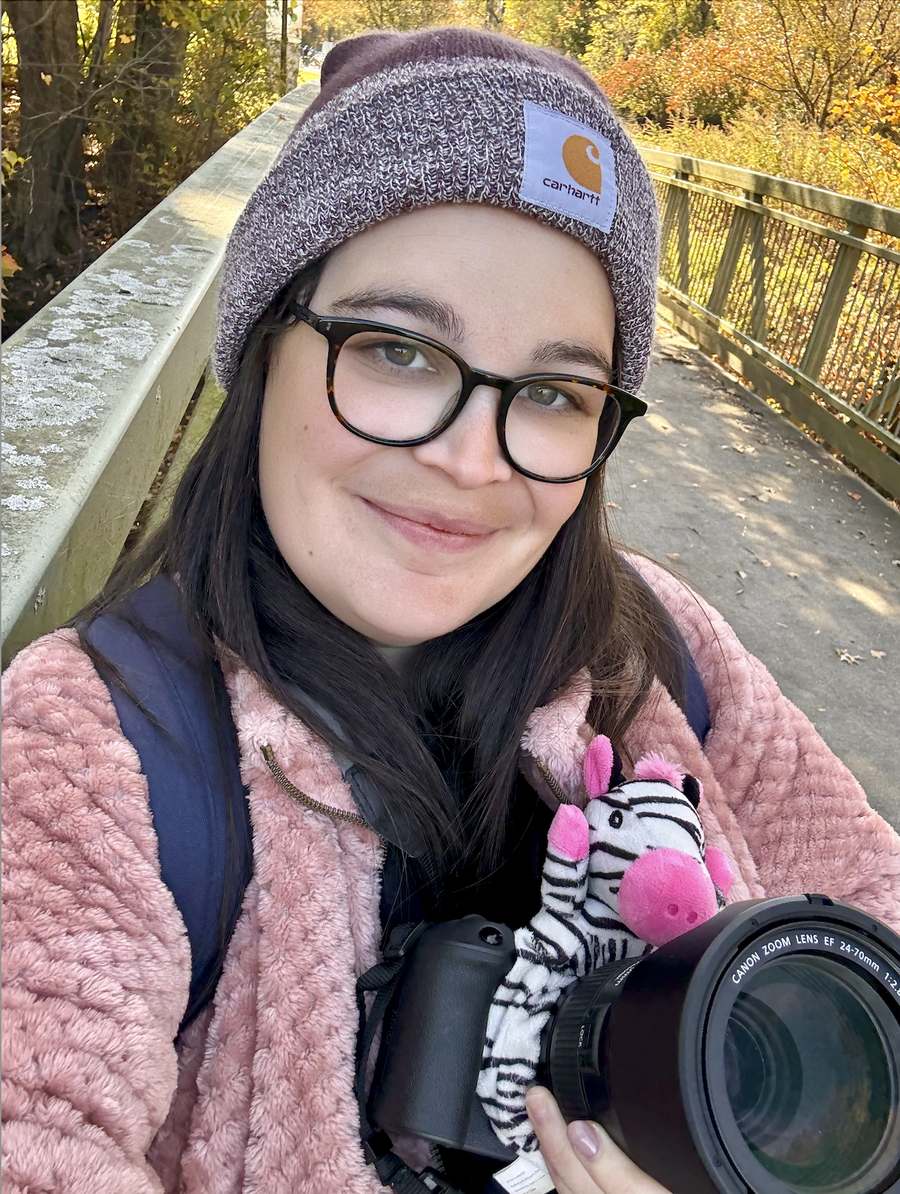 Sara Serkez in a beanie holding her camera on a bridge in autumn.