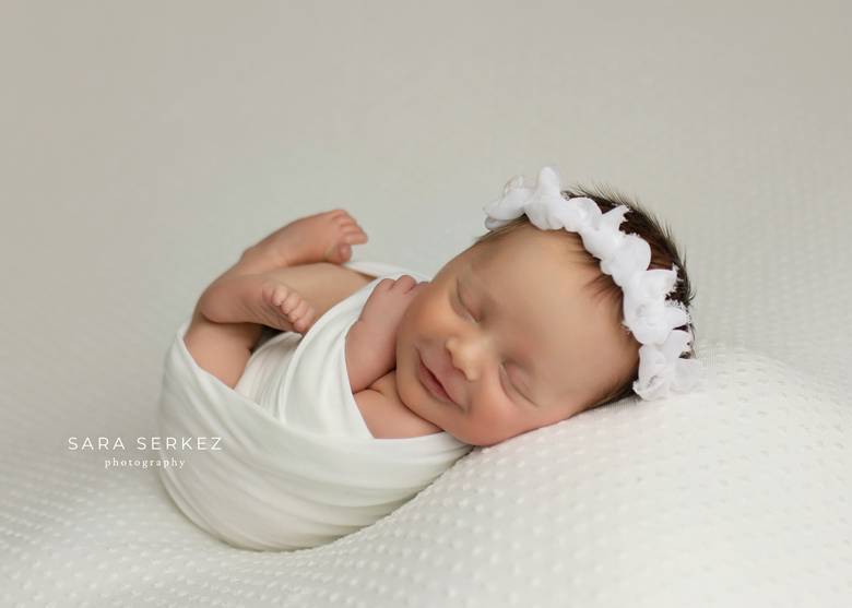 Swaddled newborn asleep and smiling on a white blanket.